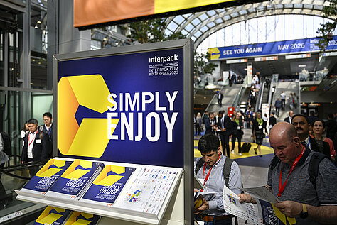 A trade fair booth of Interpack Alliance with a large blue and yellow sign saying ‘SIMPLY ENJOY’. Flyers with the logo are displayed on a stand, and visitors are visible in the background at the event.
