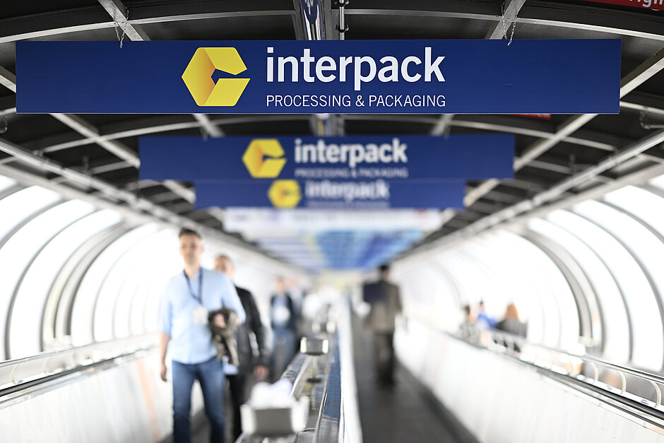People walking on a moving walkway under multiple blue signs with the yellow interpack logo and the text ‘interpack PROCESSING & PACKAGING’ in a tunnel-like structure.