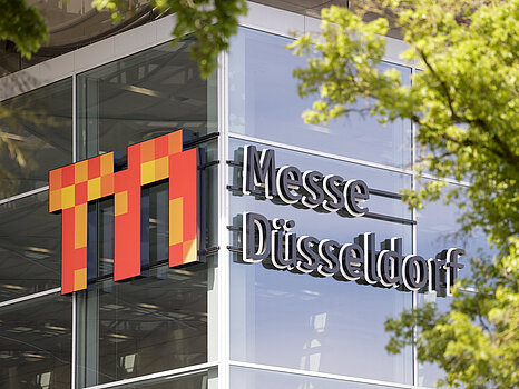 The exterior of a modern glass building with the logo ‘Messe Düsseldorf’ prominently displayed on the facade, partially framed by green tree branches.