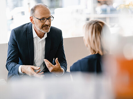 A man in a dark suit and white shirt, wearing glasses, engaging in a conversation with a woman whose back is to the camera.
